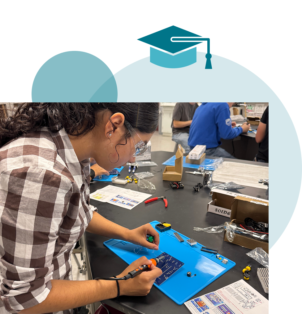 A high school student wearing safety glasses uses a soldering iron on a circuit board at a workbench, with classmates working in the background and a graduation cap icon overhead.
